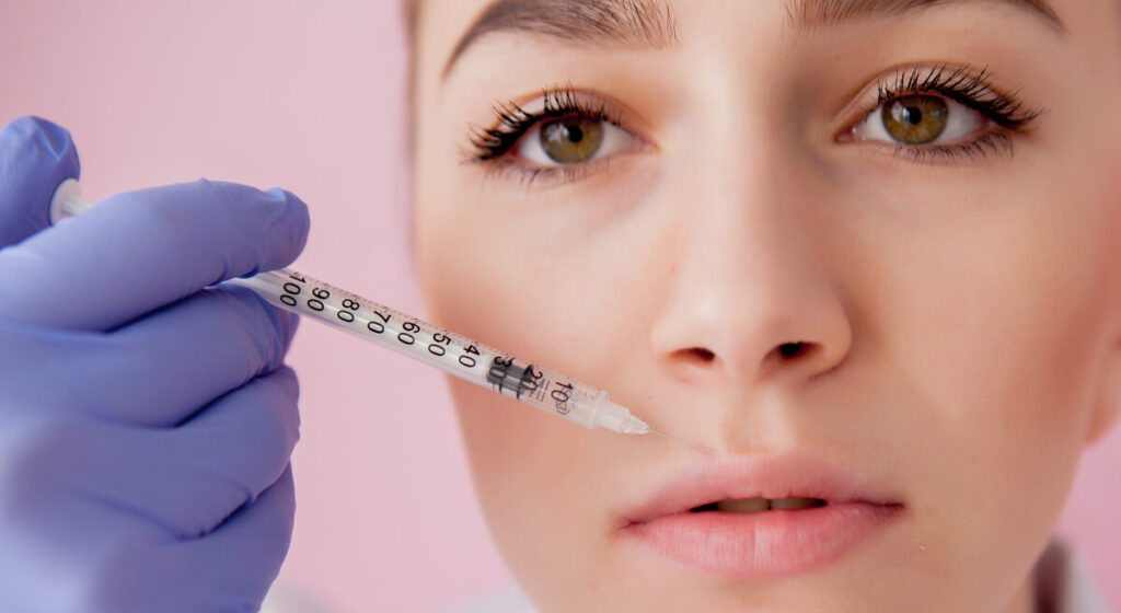 Doctor in gloves giving woman botox injections in lips, on pink background.